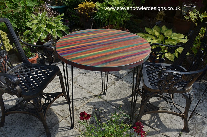 May include: A round wooden table with a colourful striped top and black hairpin legs. The table is set up outdoors with two black metal chairs.