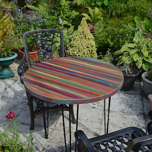 May include: A round, multi-coloured table with black hairpin legs sits on a patio with two black metal chairs. The table top is painted with stripes of various colours, including red, orange, yellow, green, blue, purple, and brown.