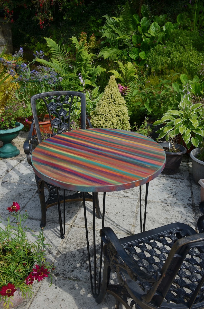 May include: A round, multi-coloured table with black hairpin legs sits on a patio with two black metal chairs. The table top is painted with stripes of various colours, including red, orange, yellow, green, blue, purple, and brown.