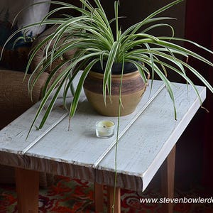 May include: A small, rustic, white-painted wooden side table with a potted spider plant. The plant has long, green and white striped leaves. A small candle in a glass holder sits on the table. The table is made of wood and has a distressed finish.