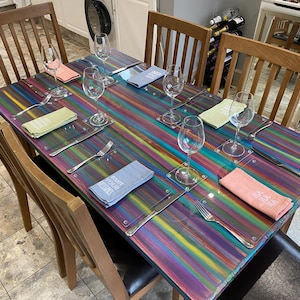 May include: A rainbow striped table with six place settings. Each place setting includes a glass, a napkin, a fork, and a knife. The table is set for a dinner party.