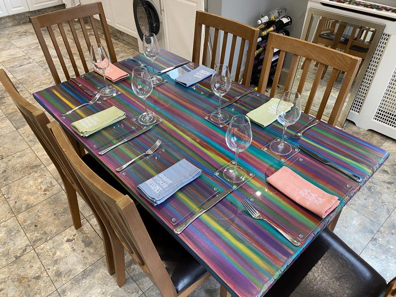 May include: A rainbow striped table with six place settings. Each place setting includes a glass, a napkin, a fork, and a knife. The table is set for a dinner party.