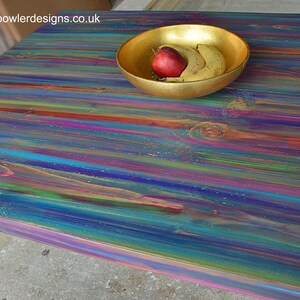 May include: A colourful wooden table top with a rainbow of colours, including blue, green, purple, and red. A gold bowl with a red apple and two bananas sits on the table.