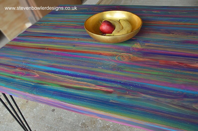 May include: A colourful wooden table top with a rainbow of colours, including blue, green, purple, and red. A gold bowl with a red apple and two bananas sits on the table.