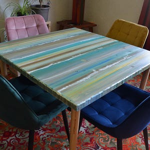 May include: A square wooden dining table with a painted top in shades of blue, green, and yellow, resembling a beach scene. The table is surrounded by four chairs, two teal and two navy blue, with wooden legs. The room has a patterned rug.