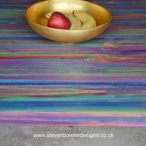 May include: A colourful wooden table top with a gold bowl filled with bananas and a red apple. The table top has a rainbow of colours, including blue, green, purple, pink, and yellow.