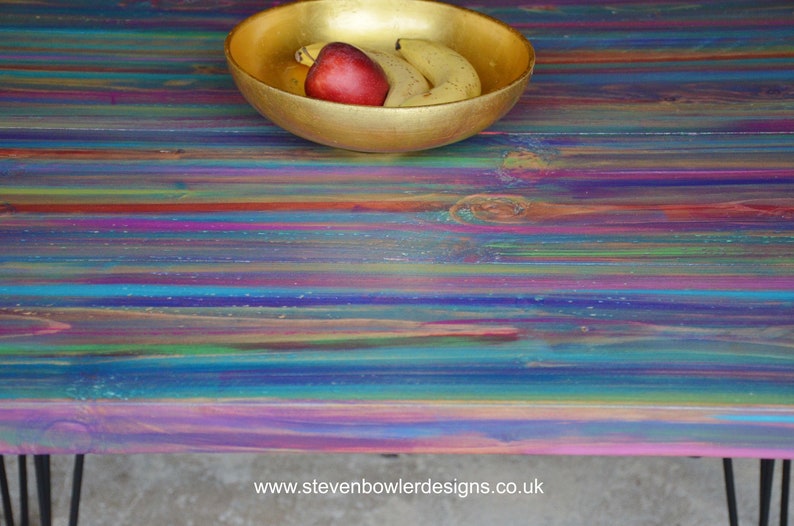 May include: A colourful wooden table top with a gold bowl filled with bananas and a red apple. The table top has a rainbow of colours, including blue, green, purple, pink, and yellow.