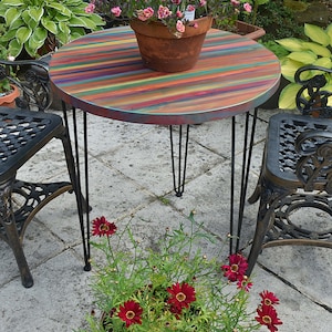 May include: A colourful striped round table with black hairpin legs, set in a garden with a potted plant and two black metal chairs.