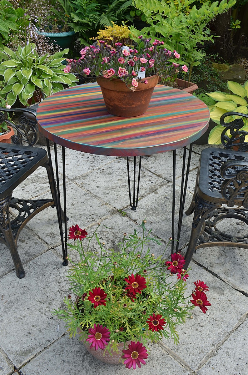 May include: A colourful striped round table with black hairpin legs, set in a garden with a potted plant and two black metal chairs.