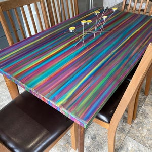May include: A wooden dining table with a colourful striped painted top. The table is surrounded by wooden chairs with brown leather seats.