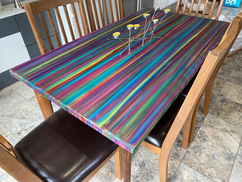May include: A wooden dining table with a colourful striped painted top. The table is surrounded by wooden chairs with brown leather seats.