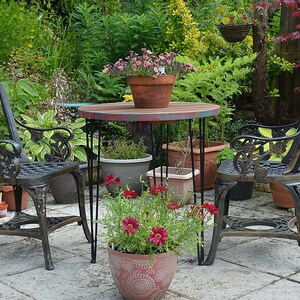 May include: A black metal patio set with two chairs and a round table. The table has a painted wooden top with a multi-coloured design. The chairs have intricate floral designs and are surrounded by potted plants and flowers.