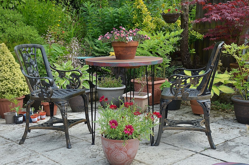 May include: A black metal patio set with two chairs and a round table. The table has a painted wooden top with a multi-coloured design. The chairs have intricate floral designs and are surrounded by potted plants and flowers.
