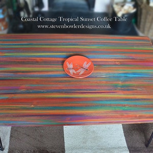 May include: A colorful wooden coffee table with a red, orange, yellow, green, blue, and purple striped top. The table has black hairpin legs and a small red plate with two white dragonflies on top.