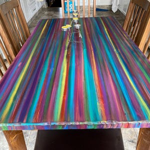 May include: A colourful wooden dining table with a rainbow striped top. The table is made from reclaimed wood and has a rustic finish. The table is surrounded by wooden chairs.