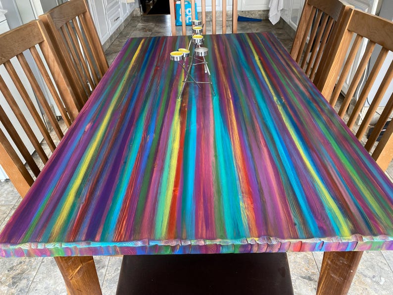 May include: A colourful wooden dining table with a rainbow striped top. The table is made from reclaimed wood and has a rustic finish. The table is surrounded by wooden chairs.