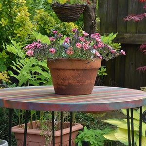 May include: A colourful striped wooden table with black metal legs, a terracotta pot with pink flowers, and a black metal garden chair in the background.