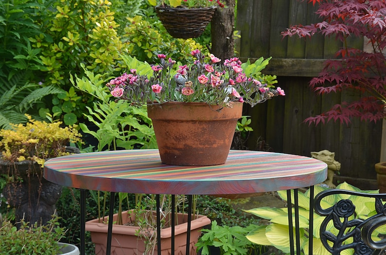 May include: A colourful striped wooden table with black metal legs, a terracotta pot with pink flowers, and a black metal garden chair in the background.
