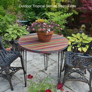 May include: A round bistro table with a colourful striped top and black hairpin legs. Two black metal chairs with ornate designs are placed on either side of the table. The table and chairs are set on a patio with a green lawn and plants in the background.