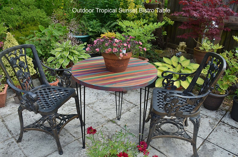 May include: A round bistro table with a colourful striped top and black hairpin legs. Two black metal chairs with ornate designs are placed on either side of the table. The table and chairs are set on a patio with a green lawn and plants in the background.