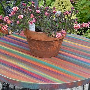 May include: A round wooden table with a colourful striped painted top. The table is outdoors and a terracotta pot with pink flowers sits on the table.