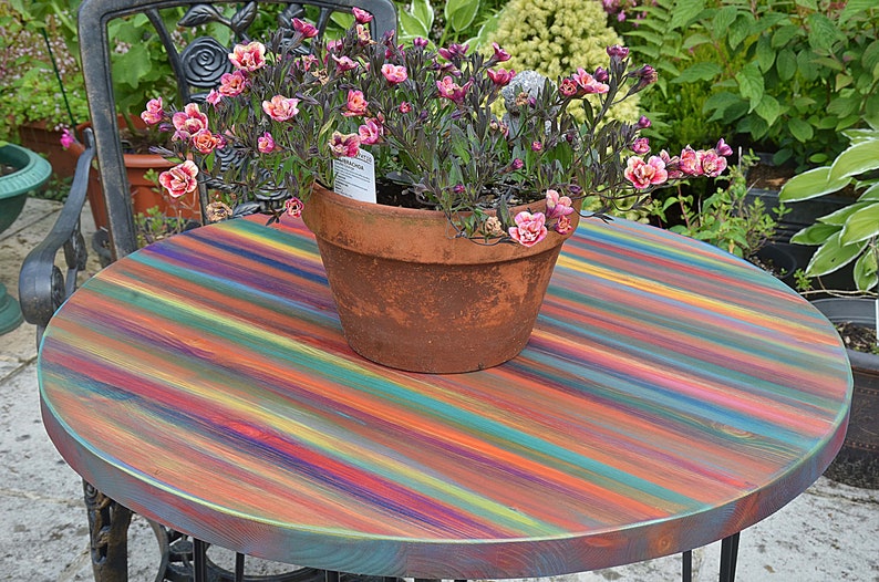 May include: A round wooden table with a colourful striped painted top. The table is outdoors and a terracotta pot with pink flowers sits on the table.