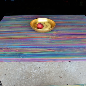 May include: A colourful wooden table with a rainbow striped finish. The table has a gold bowl with fruit on top. The table has black hairpin legs.