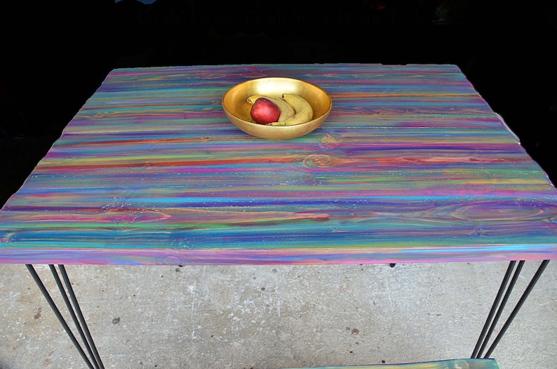May include: A colourful wooden table with a rainbow striped finish. The table has a gold bowl with fruit on top. The table has black hairpin legs.