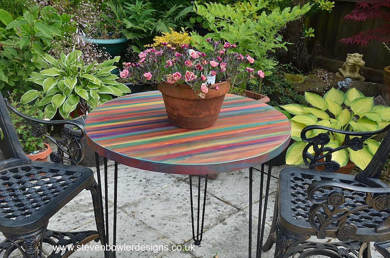 May include: A round, multi-coloured striped table with black hairpin legs sits in a garden setting. A terracotta pot with pink flowers sits on the table.