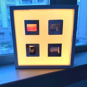 May include: A square white light box frame containing four vintage 35mm film slides arranged in a grid. The slides are backlit, displaying various outdoor scenes including a bridge, a landscape, a street view, and a horizon line over water.