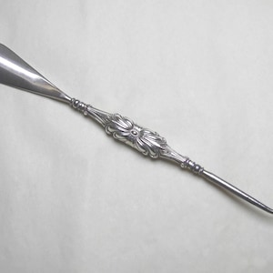 May include: A silver-toned metal shoehorn and button hook combination tool. The item features a smooth, curved scoop at one end and a small, curved hook at the opposite end. The central handle is decorated with an embossed floral pattern.