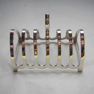 May include: A silver-coloured toast rack with six slots. The rack has a rectangular base and a handle at the top. The edges of the slots have a gold-coloured accent.