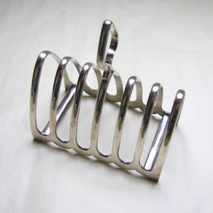 May include: A silver-toned metal toast rack featuring six arched slots for holding slices of bread. The structure is composed of curved wire loops connected by a base, with a central vertical loop at the top serving as a handle for carrying.