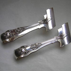 May include: A pair of silver sugar tongs, featuring decorative floral patterns on the handles. The tongs have a traditional design with curved handles and broad, flat ends for holding sugar cubes. The metal has a polished, reflective finish.