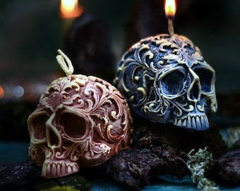 Pode incluir: Duas velas decorativas em forma de caveira com intrincados desenhos florais. As velas são feitas de cera cinza escura e marrom claro. As velas estão acesas e têm uma chama no topo.