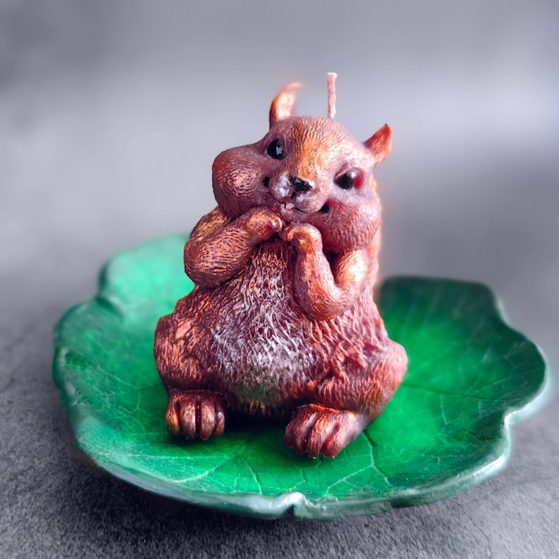 Puede incluir: Una vela de ardilla marr&oacute;n y blanca con un dise&ntilde;o detallado, sentada sobre un plato verde en forma de hoja.