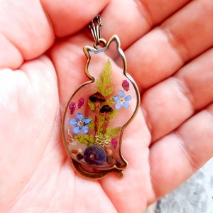 May include: A bronze cat-shaped pendant with a clear resin insert containing pressed flowers, mushrooms, and a seashell.