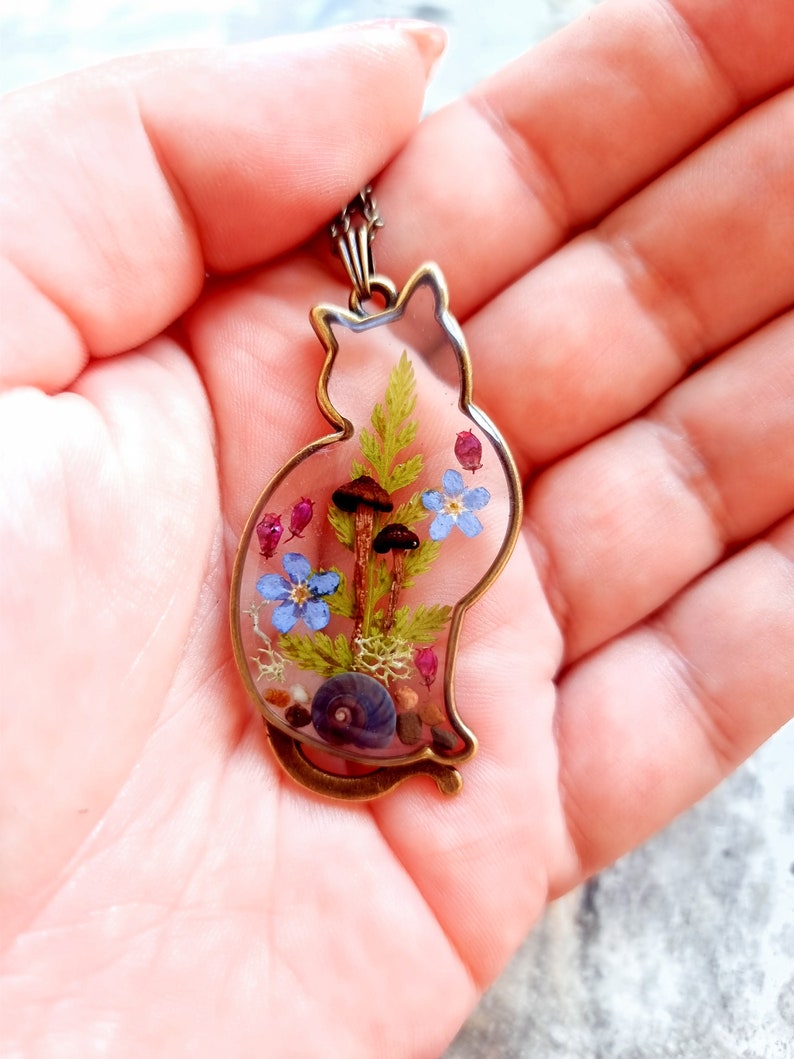 May include: A bronze cat-shaped pendant with a clear resin insert containing pressed flowers, mushrooms, and a seashell.