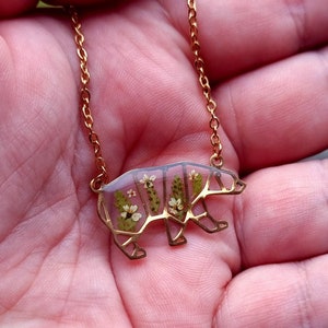 May include: A gold chain necklace with a bear-shaped pendant. The pendant is made of clear resin with pressed flowers inside.