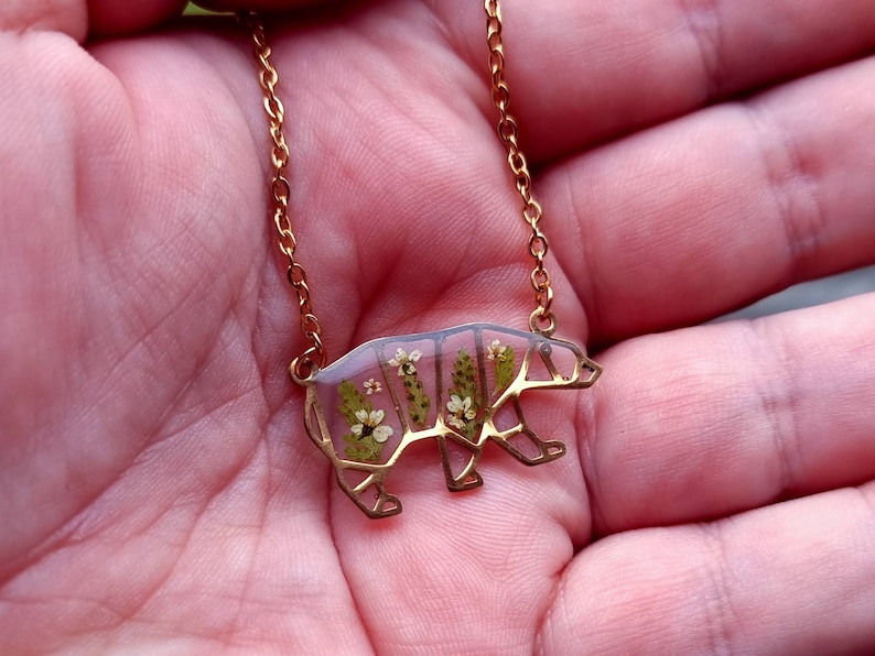 May include: A gold chain necklace with a bear-shaped pendant. The pendant is made of clear resin with pressed flowers inside.