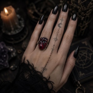 Könnte beinhalten: Ein silberner Ring mit einem detaillierten, rot geformten Herzdesign. Der Ring wird an einer Hand mit schwarzem Nagellack und einem Tattoo aus Sternen und einem Halbmond getragen. Die Gesamtästhetik ist gotisch und düster.