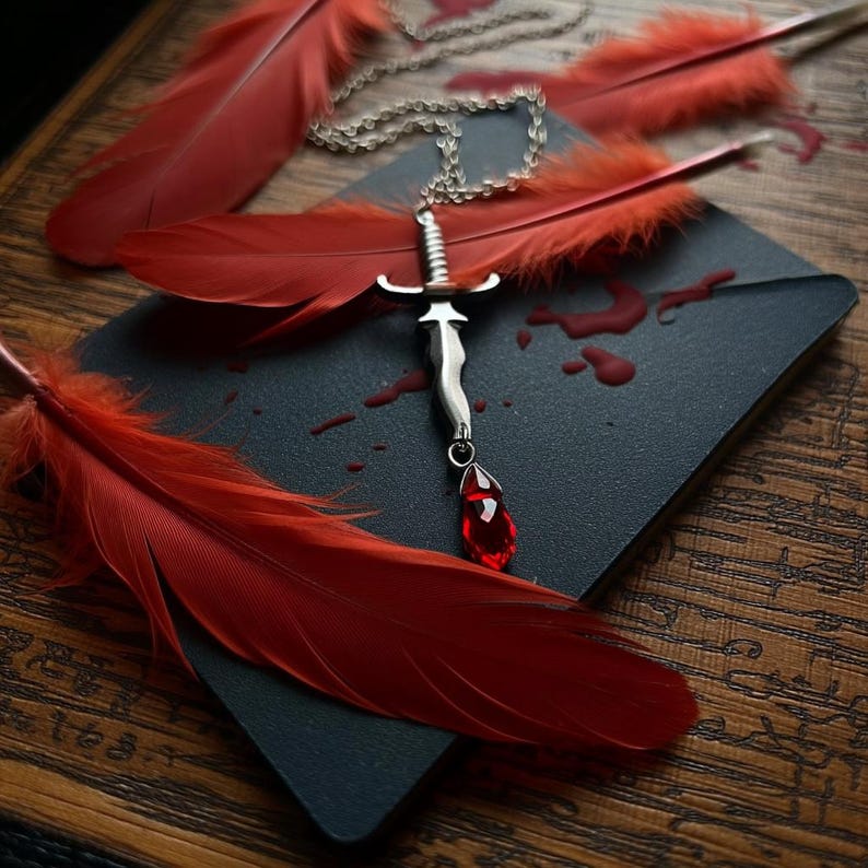 May include: A silver sword pendant with a red teardrop-shaped gem hangs from a chain. The pendant rests on a dark surface, surrounded by red feathers and simulated blood droplets. The background is a wooden surface.