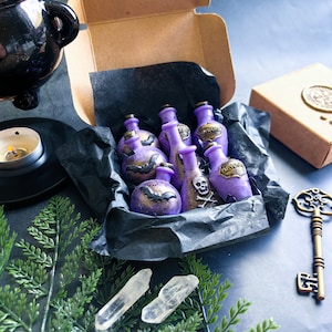 Könnte beinhalten: Ein Set aus sechs violetten Glas-Tränkflaschen mit goldenen Akzenten und Etiketten mit der Aufschrift "Beware". Die Flaschen befinden sich in einer schwarzen Geschenkbox mit Seidenpapier.