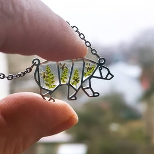 May include: A silver necklace with a bear-shaped pendant. The pendant is filled with clear resin and contains pressed green leaves and white flowers.