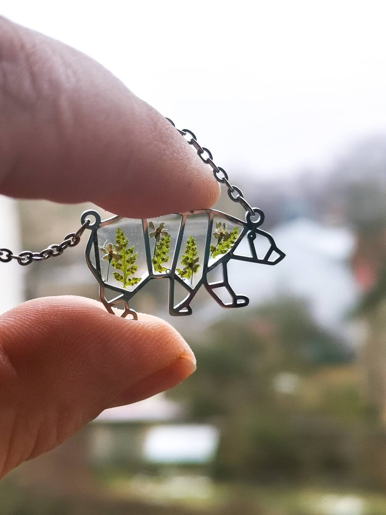May include: A silver necklace with a bear-shaped pendant. The pendant is filled with clear resin and contains pressed green leaves and white flowers.