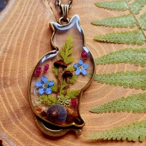 May include: A bronze cat-shaped pendant with a clear resin insert containing pressed flowers, mushrooms, and a snail shell.