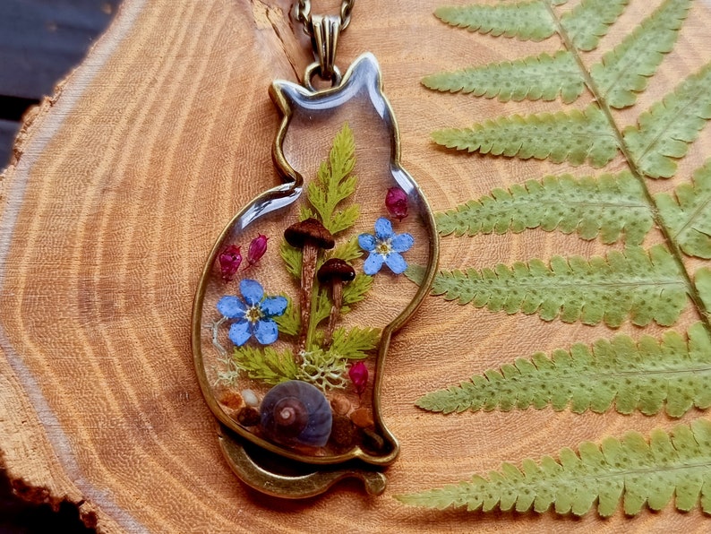 May include: A bronze cat-shaped pendant with a clear resin insert containing pressed flowers, mushrooms, and a snail shell.