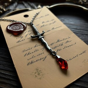 May include: A silver-toned dagger pendant necklace with a red gemstone drop. The dagger is detailed with a ribbed handle and crossguard. The necklace rests on a vintage paper with handwritten script and a wax seal.