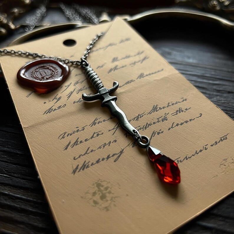 May include: A silver-toned dagger pendant necklace with a red gemstone drop. The dagger is detailed with a ribbed handle and crossguard. The necklace rests on a vintage paper with handwritten script and a wax seal.