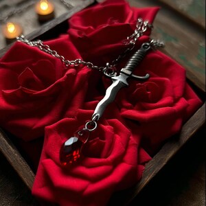 May include: A silver dagger pendant necklace with a red teardrop-shaped gem, resting on a bed of red roses. The dagger has a detailed handle and a chain. The roses are in a wooden tray.
